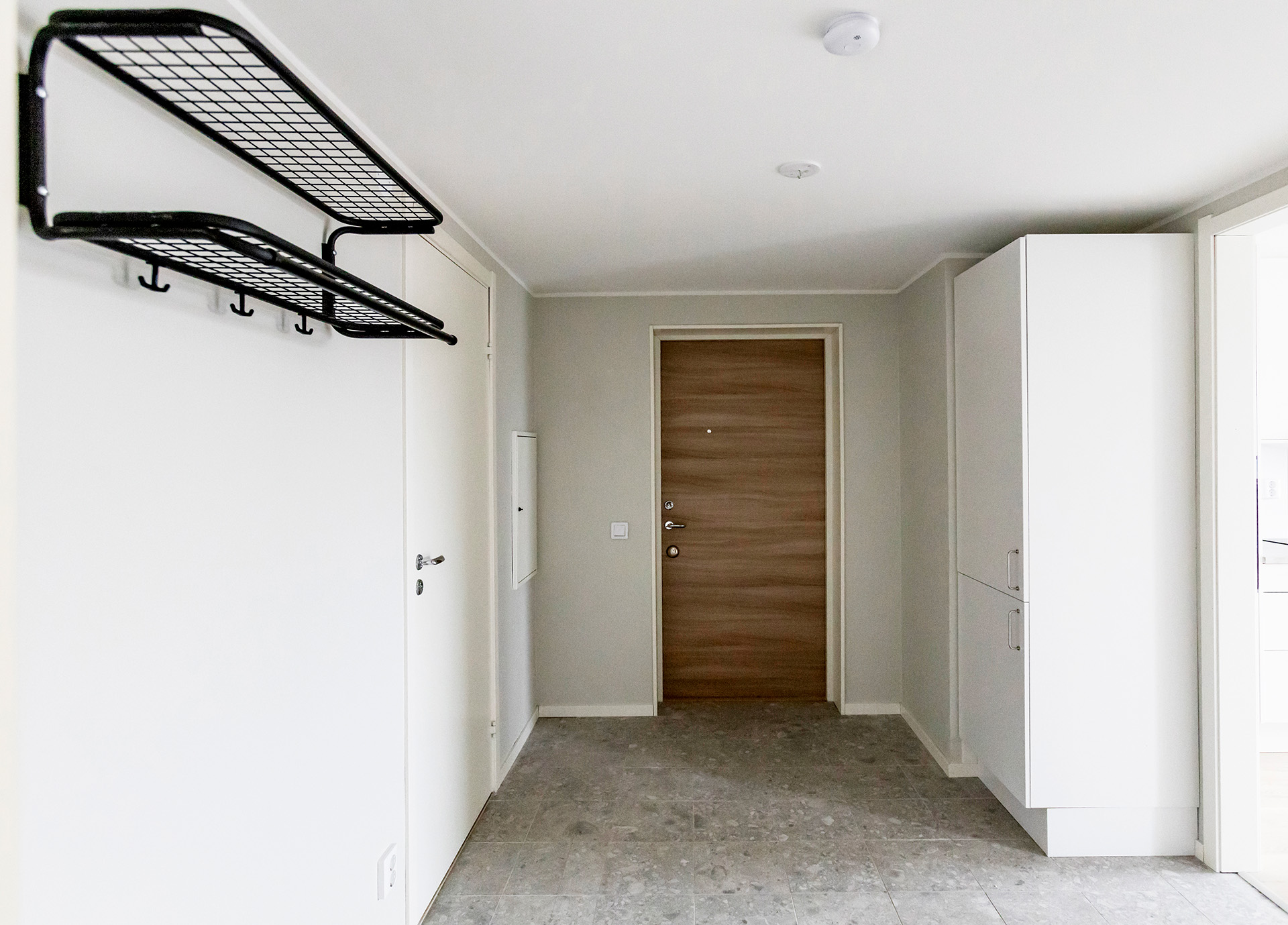 Fotograferat från sovrummet ut mot hallen. Till höger anar vi köket. Bredvid öppningen mot köket finns ett skåp. Lägenhetsdörren är i ljus ek med liggande mönster. Till vänster ser vi dörren in till badrummet och bredvid dörren finns en hatthylla. Ytskikten är ljusa: ljusgrå väggar och grått stenmönstrat klinkergolv.